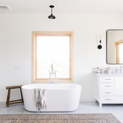 large bathroom with freestanding white tub, glass shower with large marble tile, rectangular window, black pendant light fixture, white vanity with marble top, rectangular mirror with black trim, two black sconce light fixtures, brown area rug