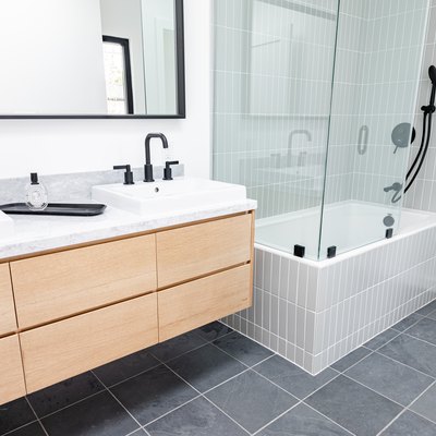 light wood floating vanity with two ceramic vessel sinks, black faucets, rectangular mirror, gray square tile floor, white tub with glass shower doors