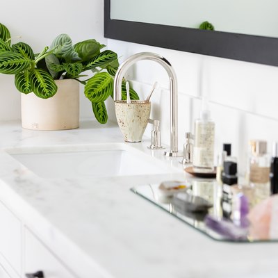 white bathroom vanity with quartz counters, white ceramic sink, silver faucet, glass tray with various beauty products, vase with green plant, rectangular mirror