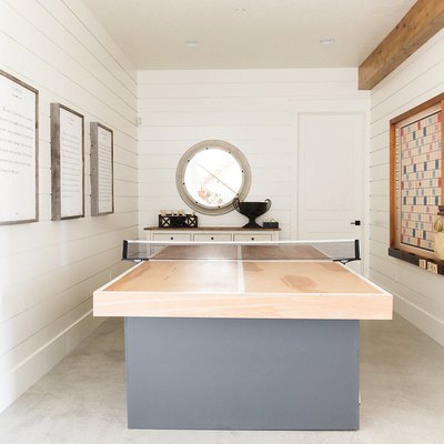 attic game room with light wood and gray ping pong table, concrete floors, white walls, round window.