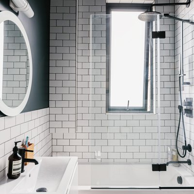small white sink, round mirror with overhead lighting, black hexagon tile on the bathtub, white subway tile on the wall, deep bathtub with a black faucet and showerhead