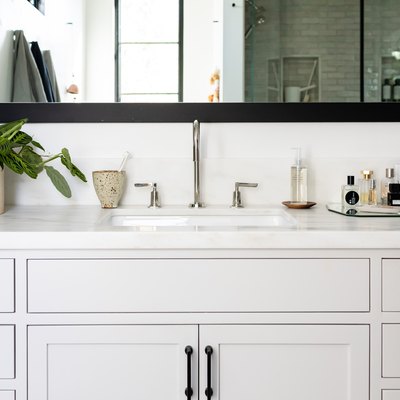 gray bathroom vanity cabinet, undermount sink, silver faucet and handles, rectangular mirror with black trim, vase with green plant, mirrored tray with various bathroom products