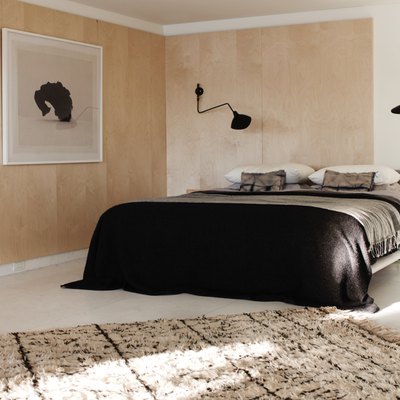 Low platform bed with black and grey linens, black modern sconces black and white large scale photo prints, natural area rug, leather side chair, wooden stool side table, flowers in vase white floors and light wood paneling on walls. Basement Bedroom Ideas