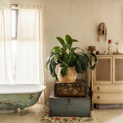 freestanding cast iron tub with vintage distress, trunks stacked on top of each other with a plant on top, a white vintage cabinet is placed between two windows