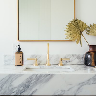 marble vanity countertop with ceramic undermount sink, gold faucet and handles, brown soap dispenser, vase with paper flowers, rectangular mirror with gold trim