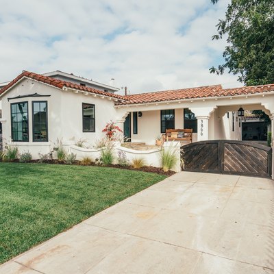 White home with Spanish-style roof, green grass lawn, concrete driveway with wooden gate