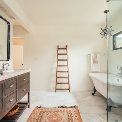 A spacious bathroom with a wood vanity with double sinks, a freestanding clawfoot tub, a glass shower door, a colorful rug, a decorative ladder leaning against the wall