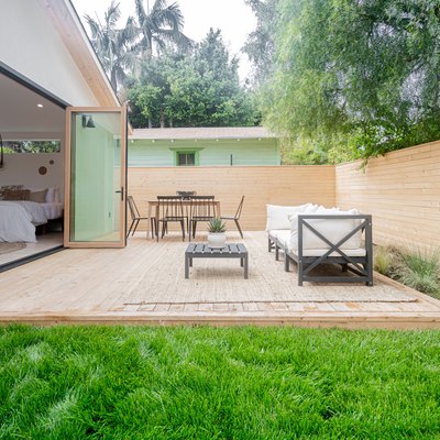 A bedroom with folding glass doors opens up to a backyard with a wood patio, wood horizontal fence, a table with five black chairs and a sofa with a coffee table; green grass and various green plants are planted