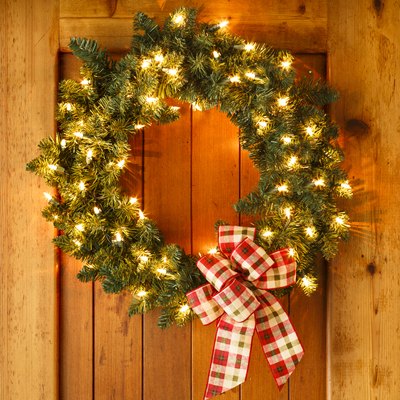 Beautiful lighted evergreen wreath with plaid fabric ribbon bow and lights hanging on wooden front door of home background. Simple, rustic country style Christmas holiday home decorations.