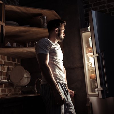 side view of bearded young man in pajamas looking at open refrigerator at night
