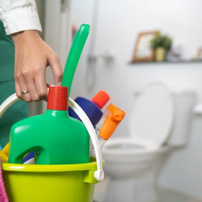 Woman standing bathroom holding a yellow plastic bucket full of bathroom cleaning supplies.