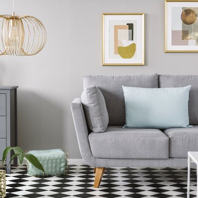 Light blue pillow placed on grey couch in bright living room interior with checkerboard linoleum floor, fresh flowers in vase on cupboard two posters hanging on the wall and gold lamp