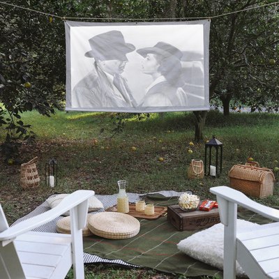 DIY portable projector screen hanging outside between two trees with chairs, blankets and snacks set in front