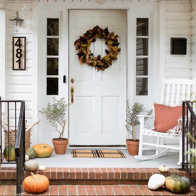 Fall porch with wreath, pumpkins, rocking chair, house numbers, and firewood stored in basket