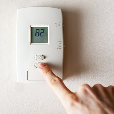 A woman is pressing the down button of a wall attached house thermostat with digital display showing the temperature