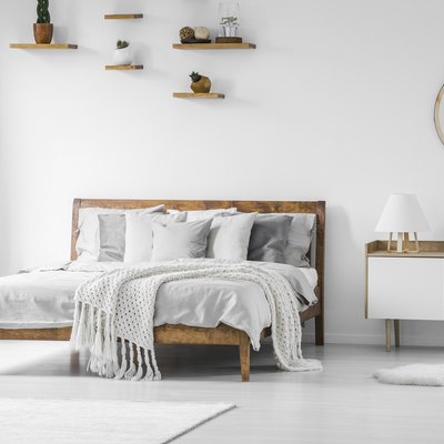 Comfortable big wooden frame bed with bed linens, pillows and blanket, nightstand beside and round mirror hanging on a white wall in a bright bedroom interior. Real photo.