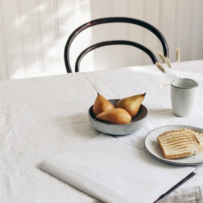 Breakfast with pears, toast, folded newspaper, and dried plants in mug on white linen table cloth.