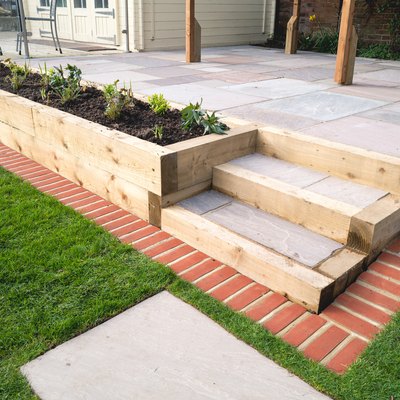New steps in a garden or back yard leading to a raised patio, alongside a new raised flowerbed made using wooden sleepers. A mowing strip of bricks is in front of newly laid turf.