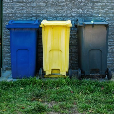 Blue, yellow, and gray trash bins standing in a row in backyard.