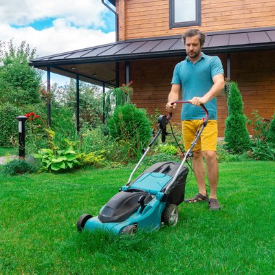 Lawn mowing with an electric lawn mower with a grass collector on a sunny summer day. A man mows the lawn in the garden of his house. An example of landscaping care.