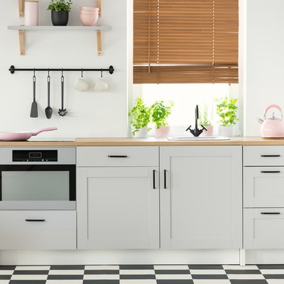 Real photo of a kitchen interior with checkered floor, white cupboards, pink accessories, plants, and window blind.