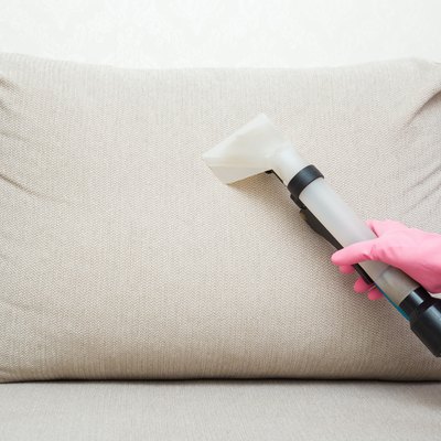 Person wearing protectives gloves using spray nozzle of professional vacuum cleaner and washing light beige back pillow of sofa at home. Extraction method. Commercial cleaning service. Closeup.
