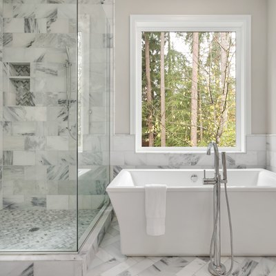 Luxury master bathroom with elegantly tiled shower in addition to a soaking bathtub installed below a large window with view of trees.