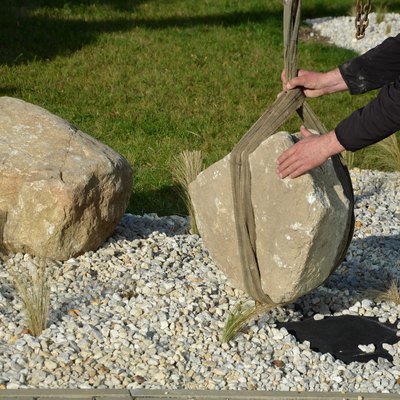 safe handling of a very heavy granite boulder with a crane, the stone is picked up and tied with straps and placed in a hole a gloved worker stabilizes the stone to the flowerbed