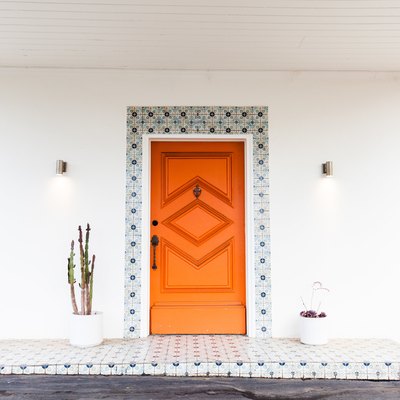 An orange front entrance door with geometric accents. Blue floral tile decorate the frame and landing. Cacti plants are on either side.