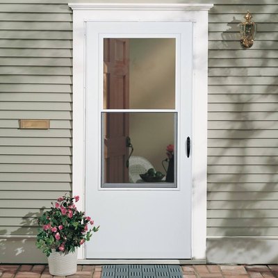 A white storm door on a house with green siding; a plant with pink flowers sits by the door