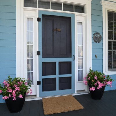 A blue screen door on a blue house with white windows; two flower pots with pink flowers are on either side of the door