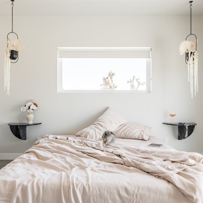 Minimalist bedroom with beige bedding, twin black shelf nightstands, double hanging planters, and rectangular window