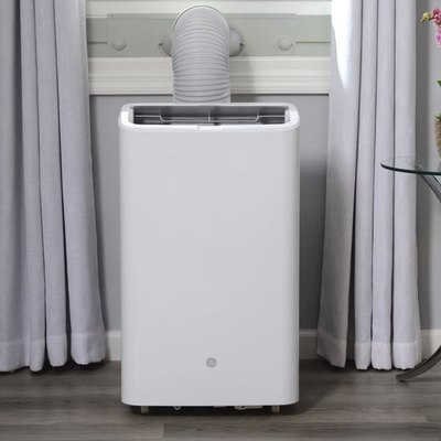 white portable air conditioner; light-gray wall and curtains, white woodwork, and a small glass/metal accent table with books and a pink potted orchid