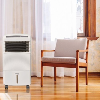White portable air conditioner in a bright corner of a room with white sheer curtains, a beige upholstered and wood chair, and polished floors