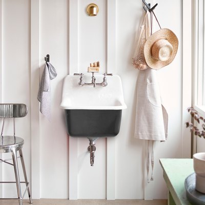 A black cast-iron wall-mounted sink on a white wall with paneling; a hat and an apron are hung near it