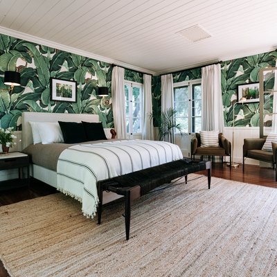 A bedroom with palm leaf wallpaper and several windows that have white curtains