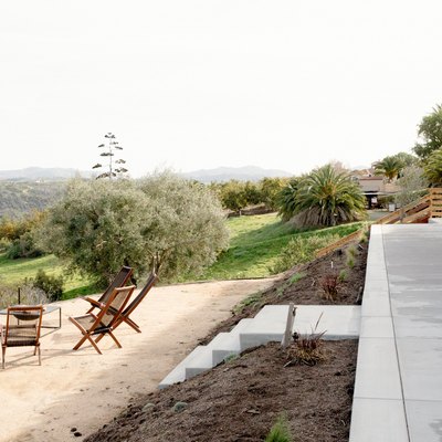 Concrete walkway with steps leading down to a dirt patio area where there are four wooden lounge chairs and a fire pit. The seating area overlooks rolling hills and a young vineyard.
