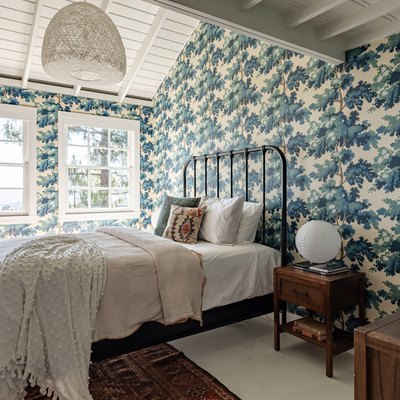Bedroom with vintage blue tree wallpaper, black bar headboard, lanterns, neutral bedding, and double windows.