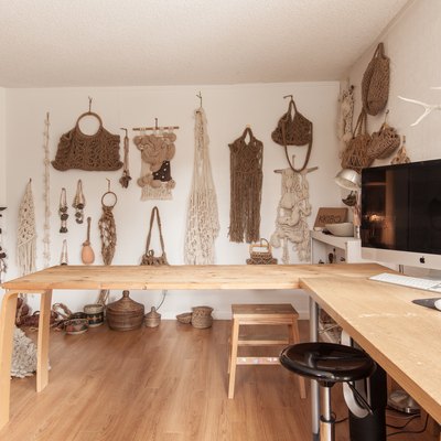 Studio with a display wall of macrame purses and wall hangings. A wood L-shape desk with a computer.