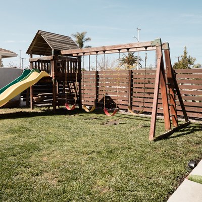 A wooden swing set on top of a patch of grass; a wooden horizontal fence and square concrete pavers