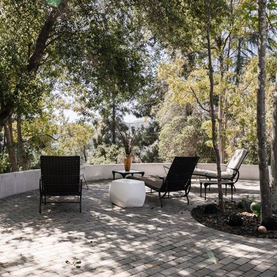 Gray brick patio with chaise lounge chairs, and a white geometric side table. Trees and plants surround.