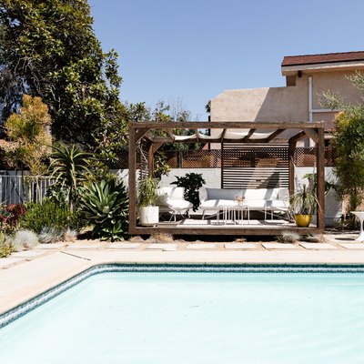 Swimming pool with a wood pergola with white patio furniture and tropical plants.