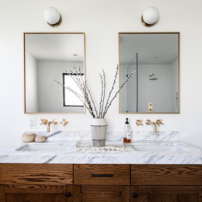 wood bathroom vanity with marble vanity top, a black and white vase with flowers, two mirrors over double sinks with white, round sconces overhead