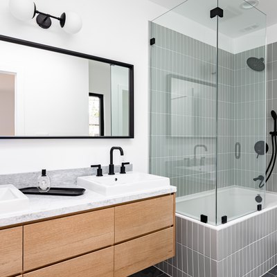 Modern bathroom with glass-walled shower over white-tiled bathtub, black faucets and shower head, white sinks, and black-framed mirror