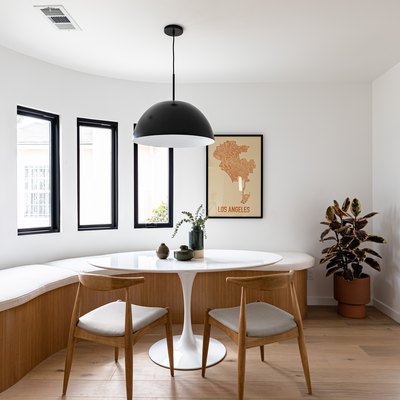 Contemporary curved dining room with curved bench, small round table, chairs, pendant lamp, framed print, and tall windows with view of hallway via arch