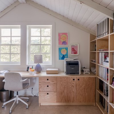 An office with a wood bookcase and Scandinavian style wood desk. A gray office chair and gray table lamp. The desk is by a window.