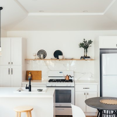 Small kitchen with white cabinets and marble backsplash. Wood open shelving, and one hanging pendant light.