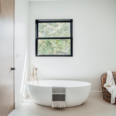 Minimalist bathroom with light wood door, white walls and floors, freestanding bathtub with towel, peacock chair with white towel