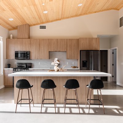 Minimalist kitchen with wood cabinets, white counters, black bar stools, and a sloped wood ceiling.