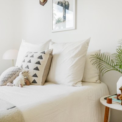 White bedding, black-white patterned pillows, stuffed animal and blanket. Stool with geometric vase of palm leaves and a sculpture